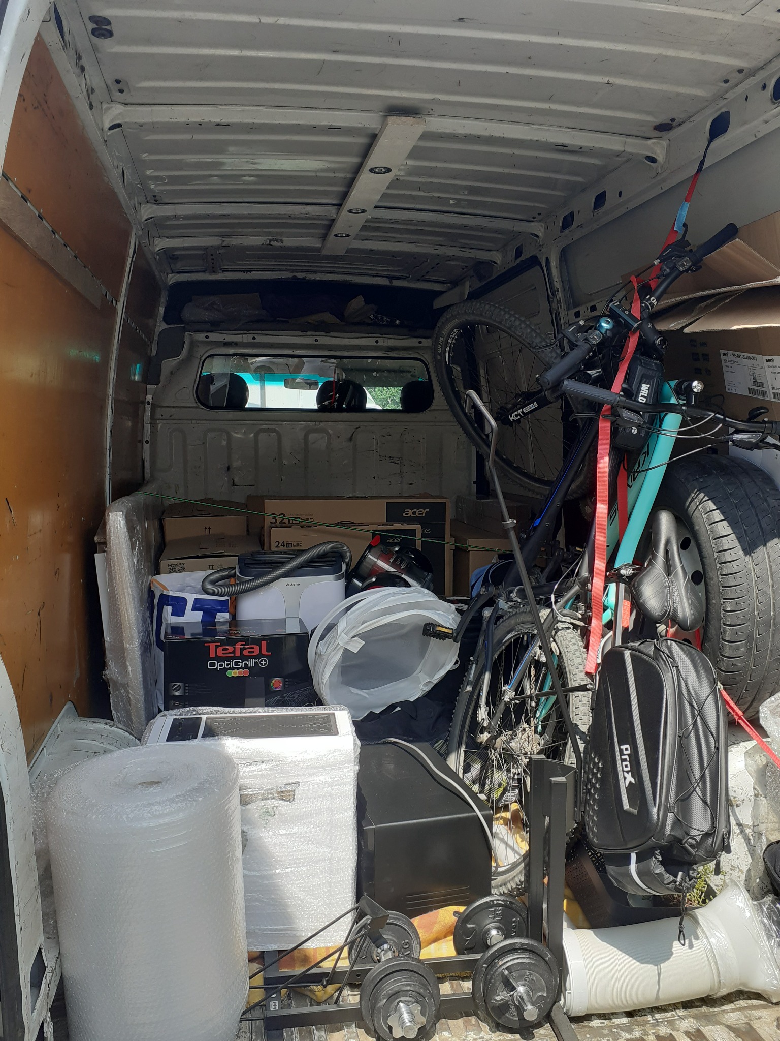 Wnętrze furgonetki załadowane różnorodnymi przedmiotami do przeprowadzki: rower, kartony, sprzęt AGD, rolka folii bąbelkowej, hantle i torba rowerowa PROX