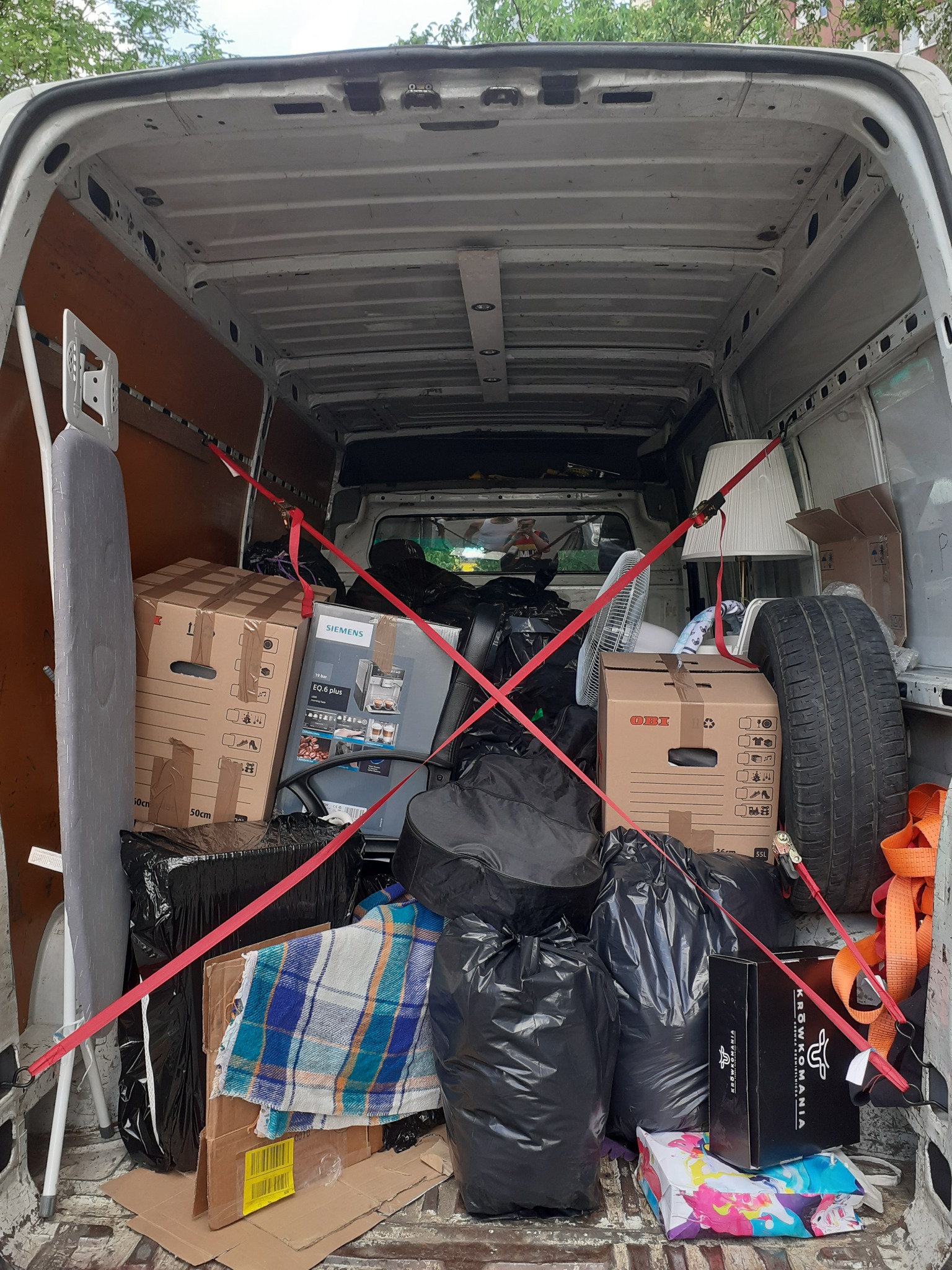 Wnętrze białego furgonetki wypełnione po brzegi różnorodnymi przedmiotami zabezpieczonymi czerwonymi pasami transportowymi, w tym kartony, torby na śmieci, deska do prasowania, wentylator, opona...