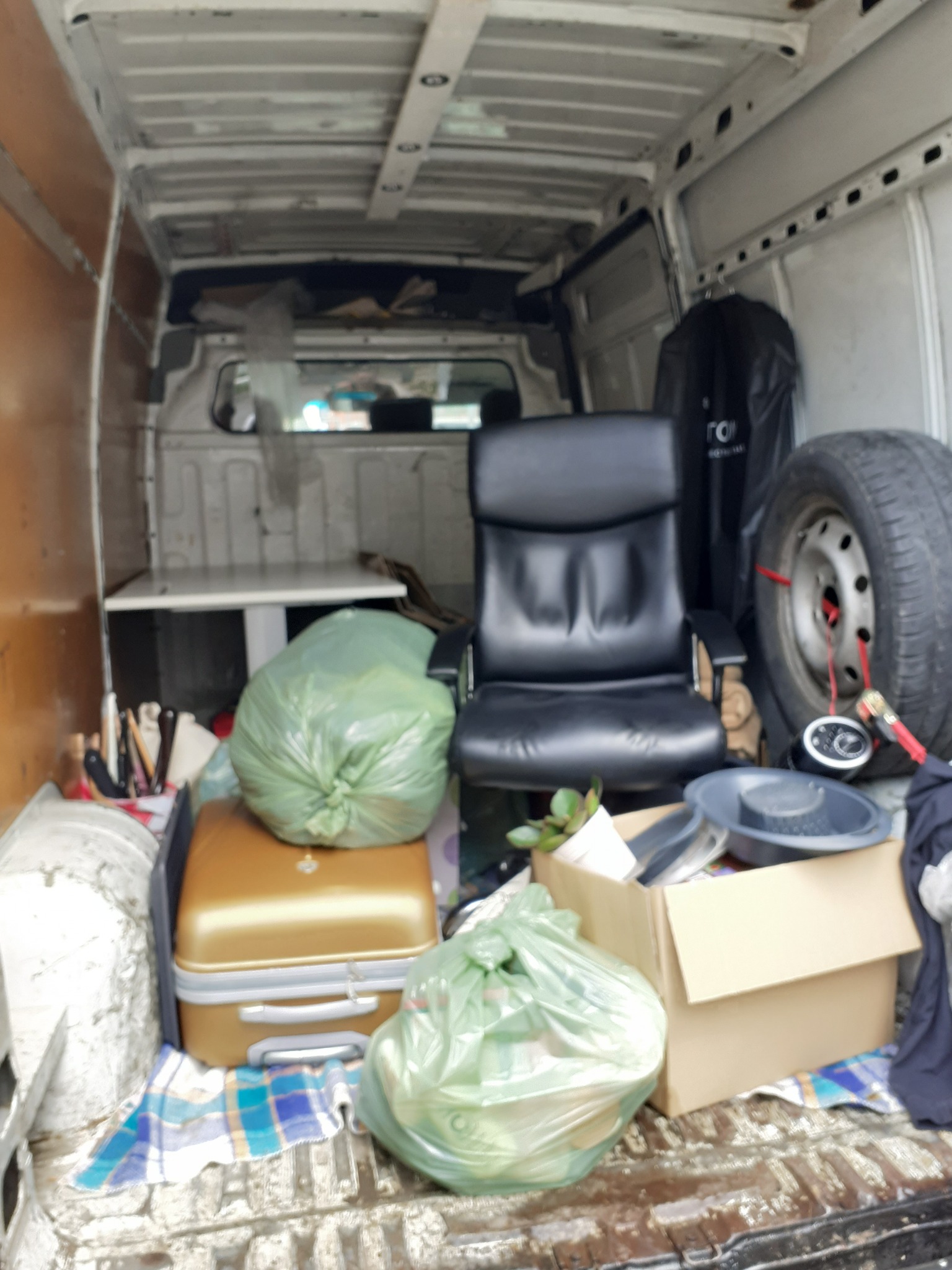 Wnętrze furgonetki załadowane różnymi przedmiotami: krzesło biurowe, torby z zawartością, walizka, koło samochodowe i inne akcesoria, widok z tyłu pojazdu.