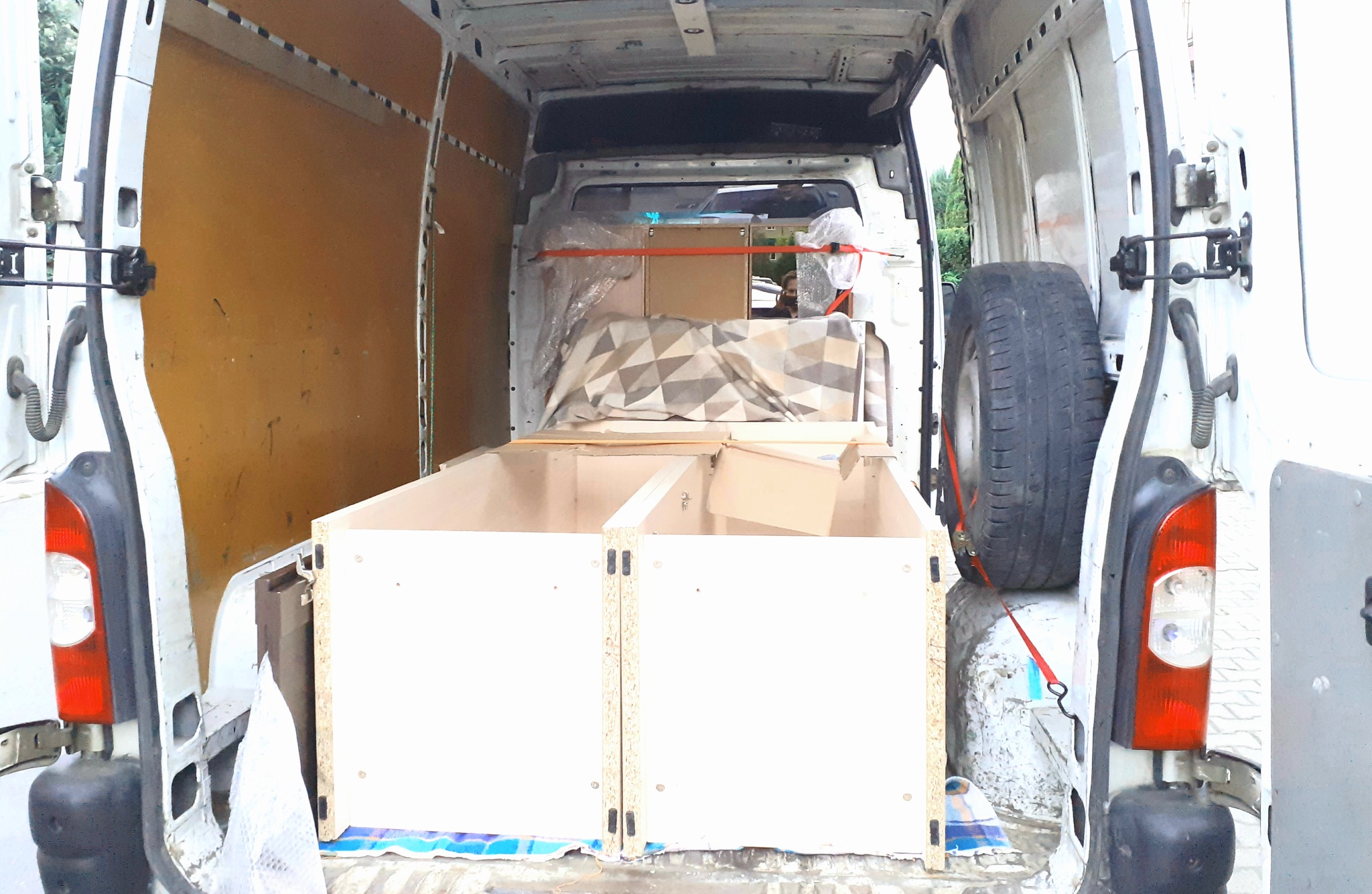 Wnętrze białego dostawczego vana z otwartymi tylnymi drzwiami, częściowo załadowane: widoczne dwie jasne, drewniane skrzynie z płyty wiórowej, zapakowane kartony, koc w geometryczne wzory, opona...