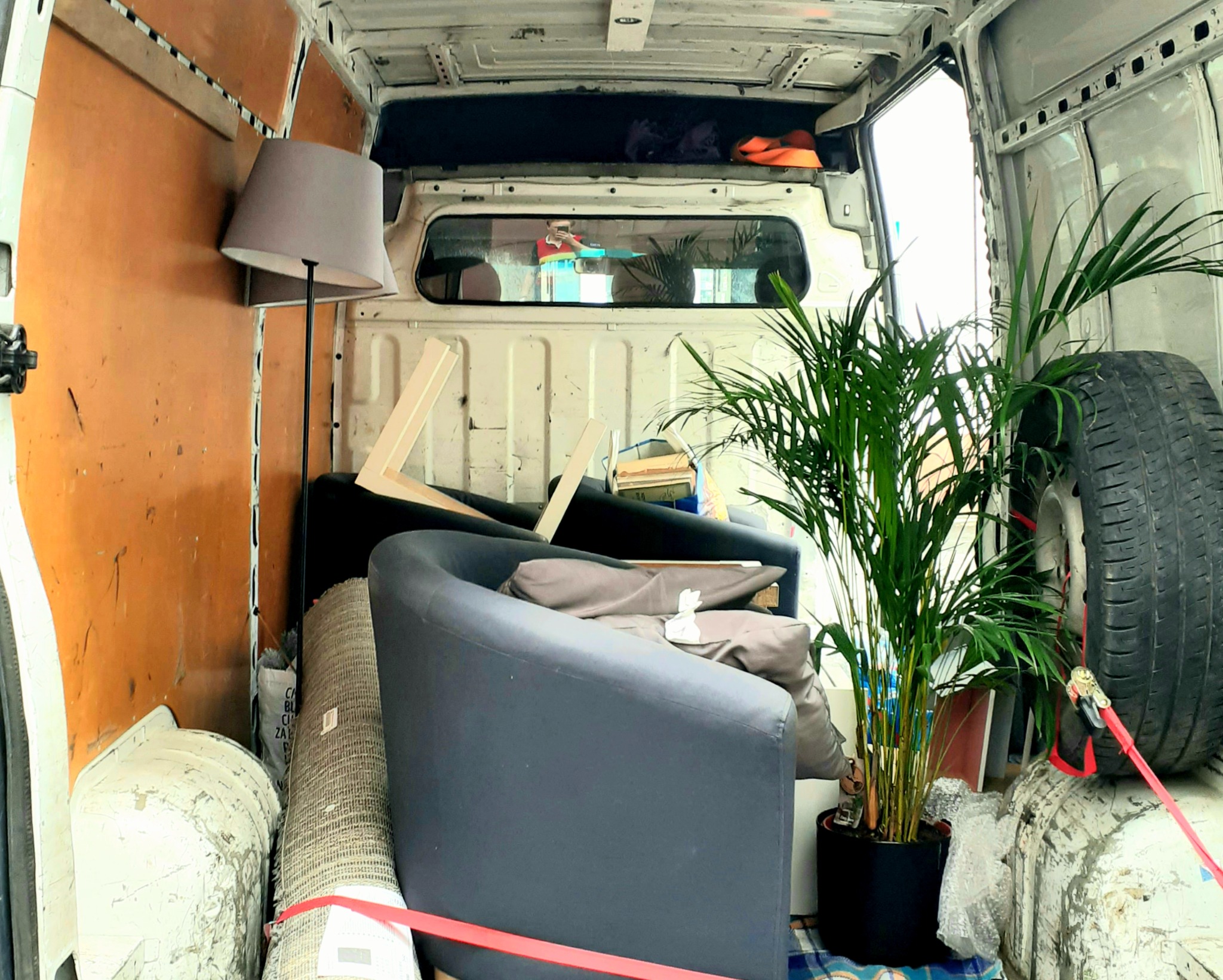 Wnętrze paki busa załadowane meblami: dwa fotele, lampa, dywan zwinięty w rulon, roślina doniczkowa i opona zabezpieczone czerwonym pasem transportowym.