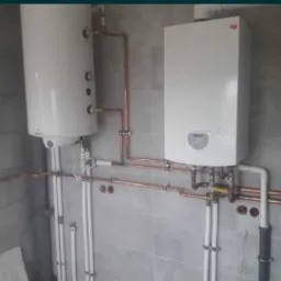 Biały bojler i piec gazowy zamontowane na szarej, kafelkowanej ścianie, połączone miedzianymi rurami. Widoczne także rury PCV.