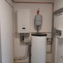 Biała pompa ciepła LG Therma V z szarym zbiornikiem wyrównawczym i zasobnikiem ciepłej wody w kotłowni z miedzianymi rurami instalacji grzewczej.