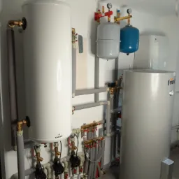 Instalacja hydrauliczna w kotłowni: białe i szare zbiorniki akumulacyjne, rury w izolacji, zawory z czerwonymi pokrętłami, manometry, widoczny panel sterowania na ścianie.