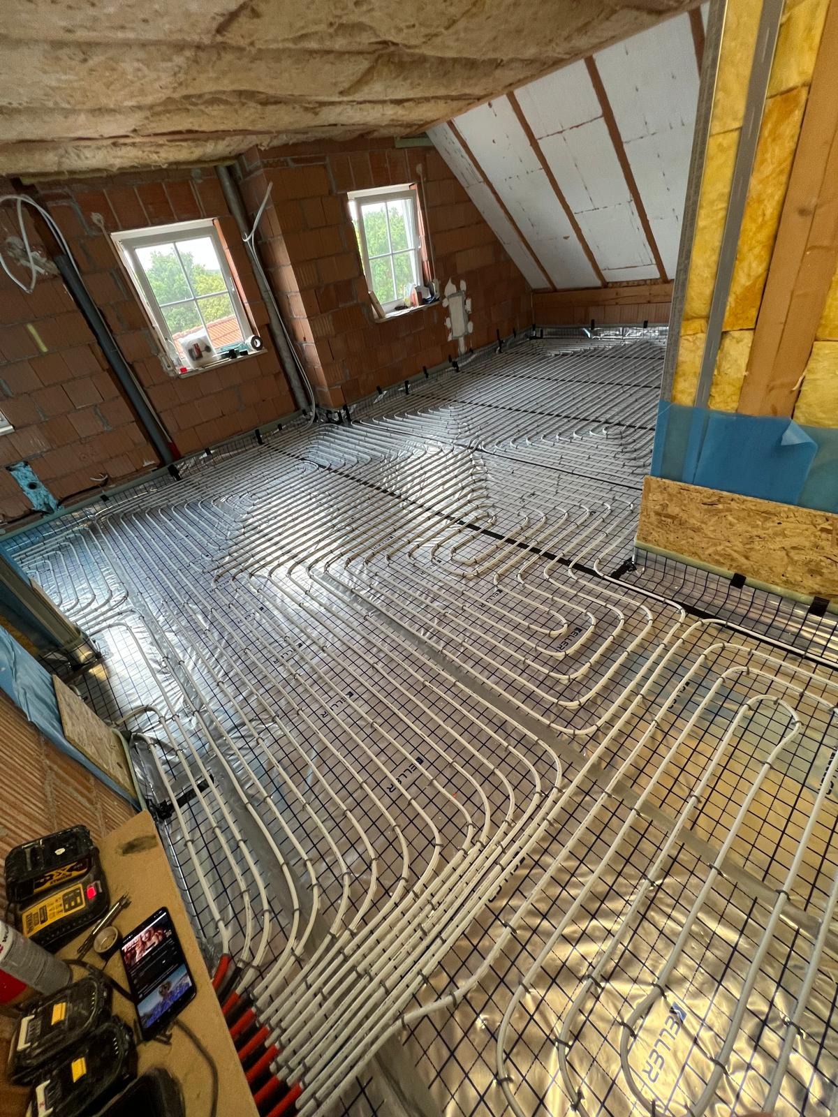 Instalacja ogrzewania podłogowego w trakcie montażu na poddaszu, widoczne rury grzewcze ułożone na folii, okna dachowe i fragmenty narzędzi pracy.