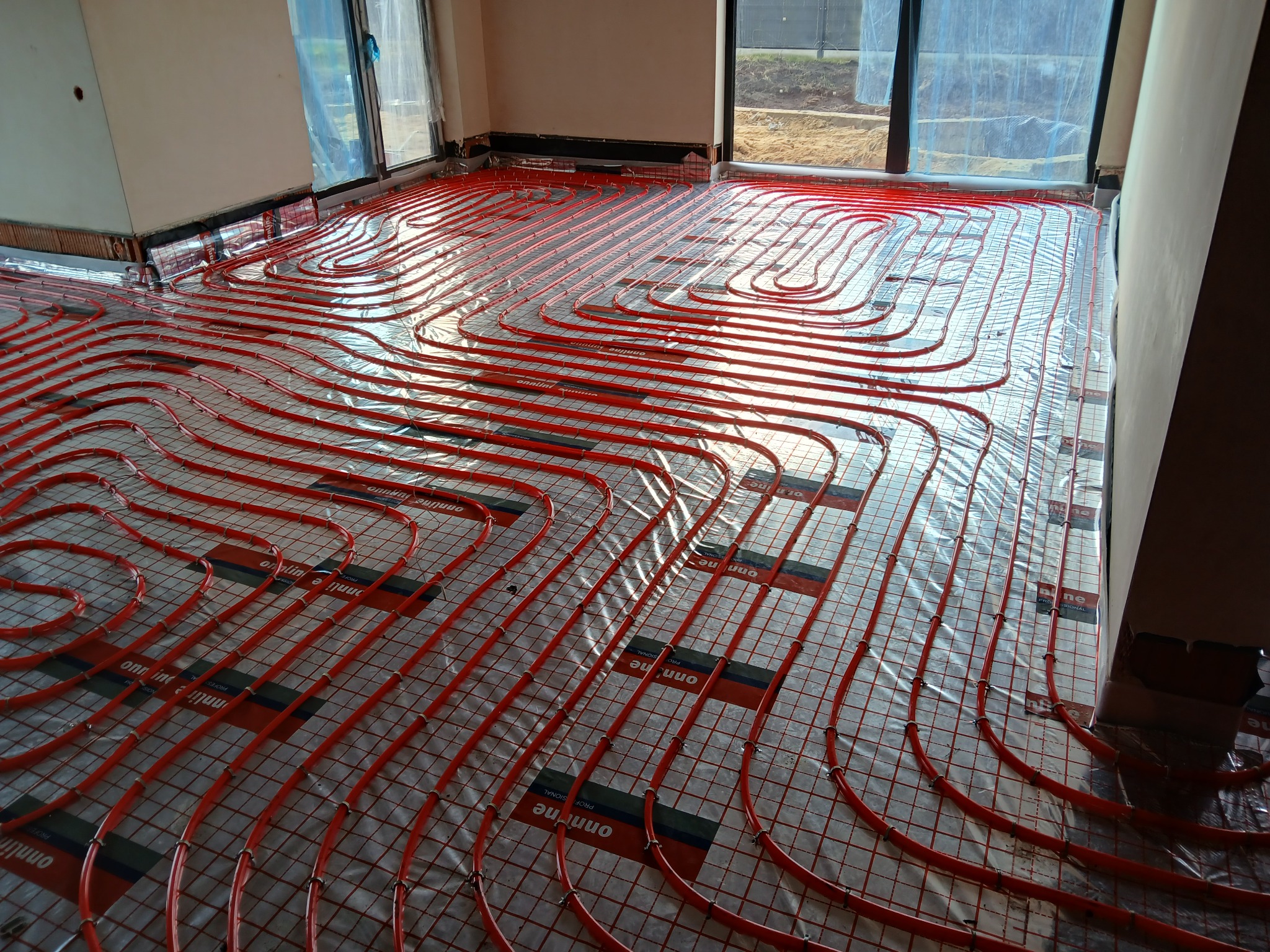 Czerwone rury ogrzewania podłogowego ułożone na folii, widoczne okna w tle. Instalacja w trakcie realizacji, przed wylaniem posadzki. Charakterystyczny wzór ułożenia rur.