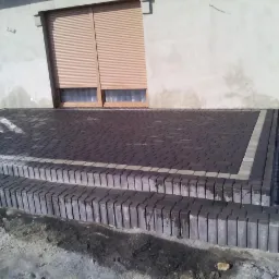 Schody i taras z ciemnej kostki brukowej z jasnoszarą obwódką przy szarym budynku z roletami, widoczne ślady zaprawy na kostce.