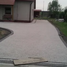 Szeroki, zakrzywiony podjazd z jasnoszarej kostki granitowej, obrzeża z ciemniejszej kostki, widok na dom z czerwonym dachem w tle, częściowo widoczny pracownik i taczka.