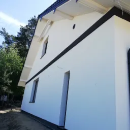 Nowo pomalowana, biała elewacja domu jednorodzinnego z czarnymi akcentami, widok z dołu, z widoczną ziemią i fragmentem zieleni w tle.