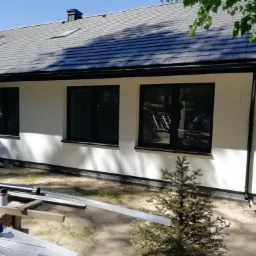 Biała elewacja domu z czarnymi ramami okiennymi, widoczne elementy rusztowania i mała choinka na pierwszym planie, odbicie osoby pracującej w oknie.