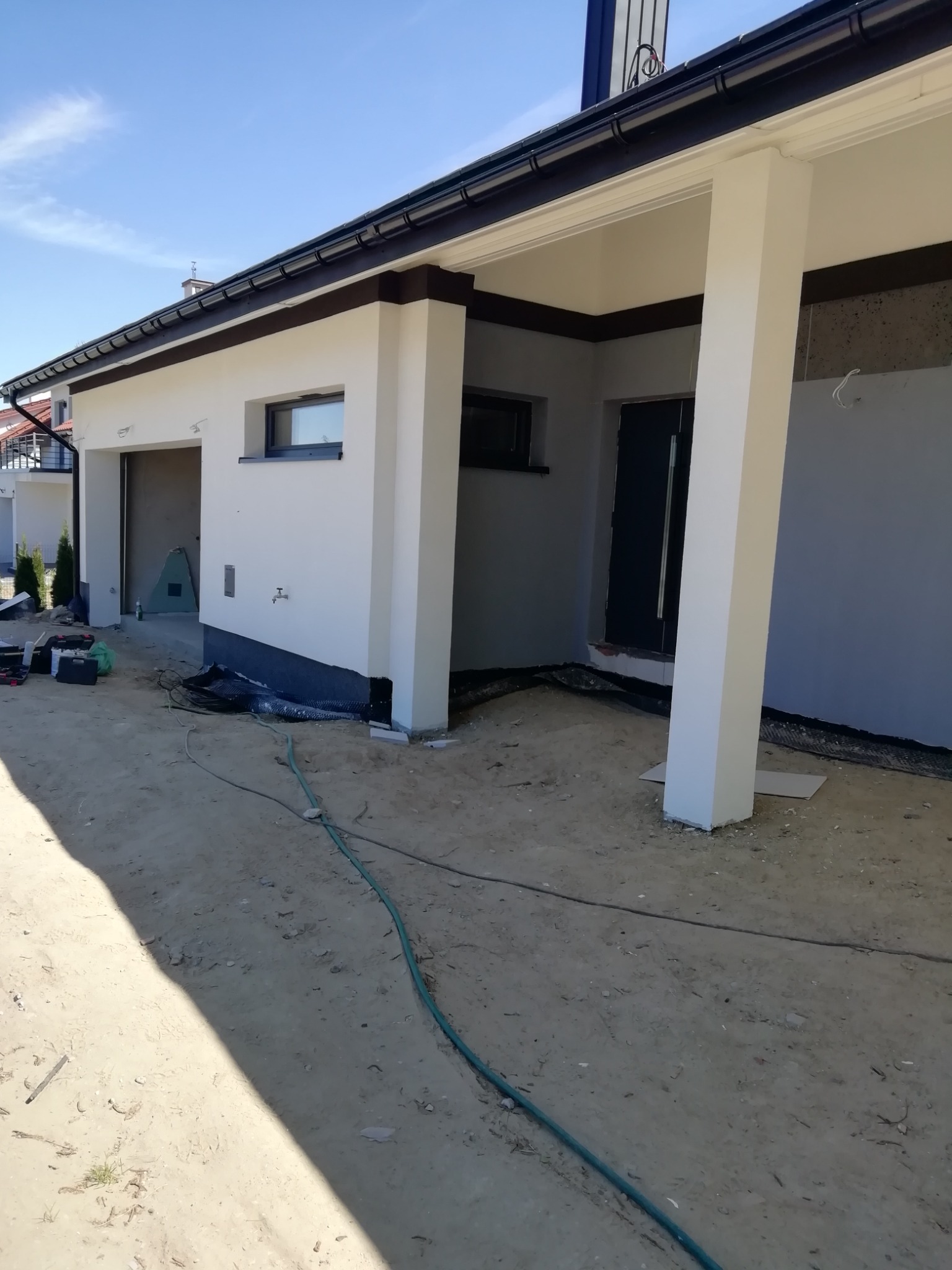 Nowoczesny dom jednorodzinny w trakcie prac wykończeniowych elewacji: widoczne fragmenty ocieplenia przy gruncie, świeżo pomalowane ściany na biało, czarne rynny i stolarka okienna, ziemia wokół...