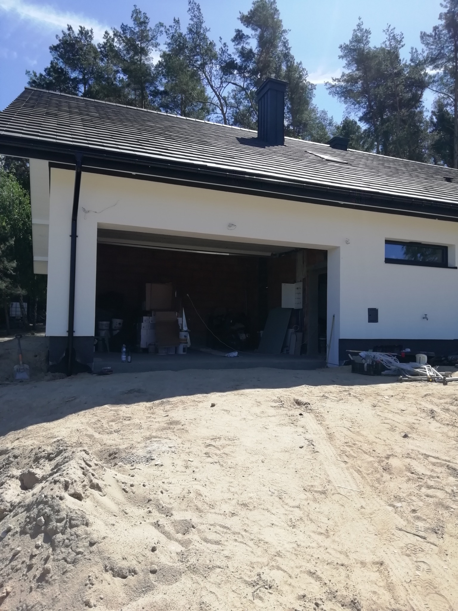 Nowoczesny budynek garażowy z białą elewacją i ciemną dachówką, widok z zewnątrz na plac budowy z piaskiem.