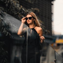 MAUPACYKA.PL Marcin Klinikowski - Elegancka blondynka w czarnym kombinezonie i okularach przeciwsłonecznych, pozująca na tle miejskiego krajobrazu z elementami zieleni i nowoczesnej architektury.