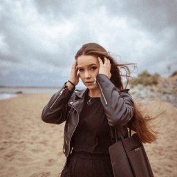 MAUPACYKA.PL Marcin Klinikowski - Portret młodej kobiety w czarnej skórzanej kurtce i sukience na plaży przy pochmurnym niebie, z rozwianymi włosami i torbą na ramieniu, patrzącej w obiektyw z rękami uniesionymi do głowy.