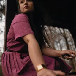 Kobieta w fioletowej sukience w kropki siedzi w altanie, spoglądając w lewo, z widocznym złotym zegarkiem i pierścionkiem na palcu.