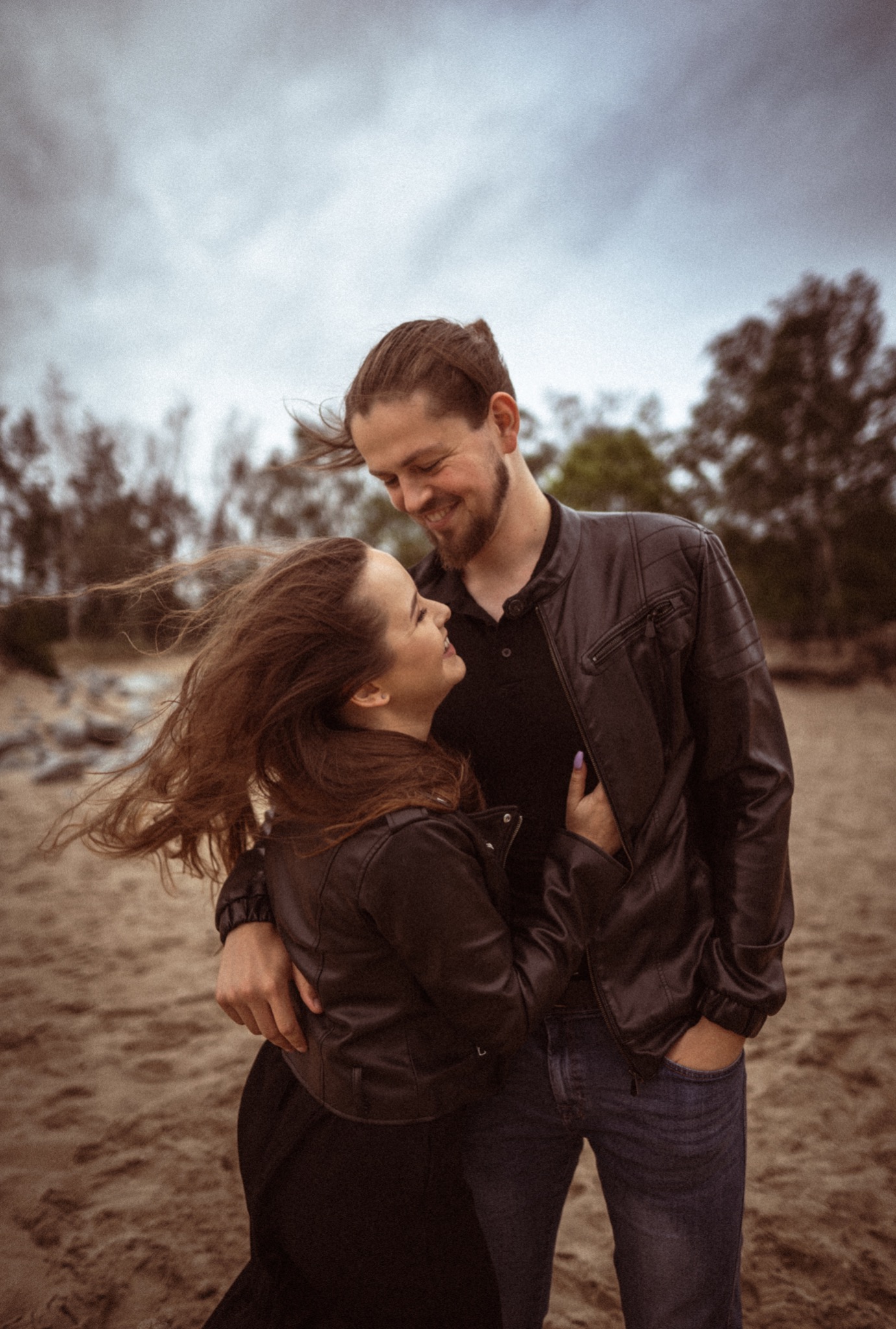 Uśmiechnięta para w skórzanych kurtkach obejmuje się na piaszczystej plaży, włosy kobiety rozwiane na wietrze, pochmurne niebo w tle.