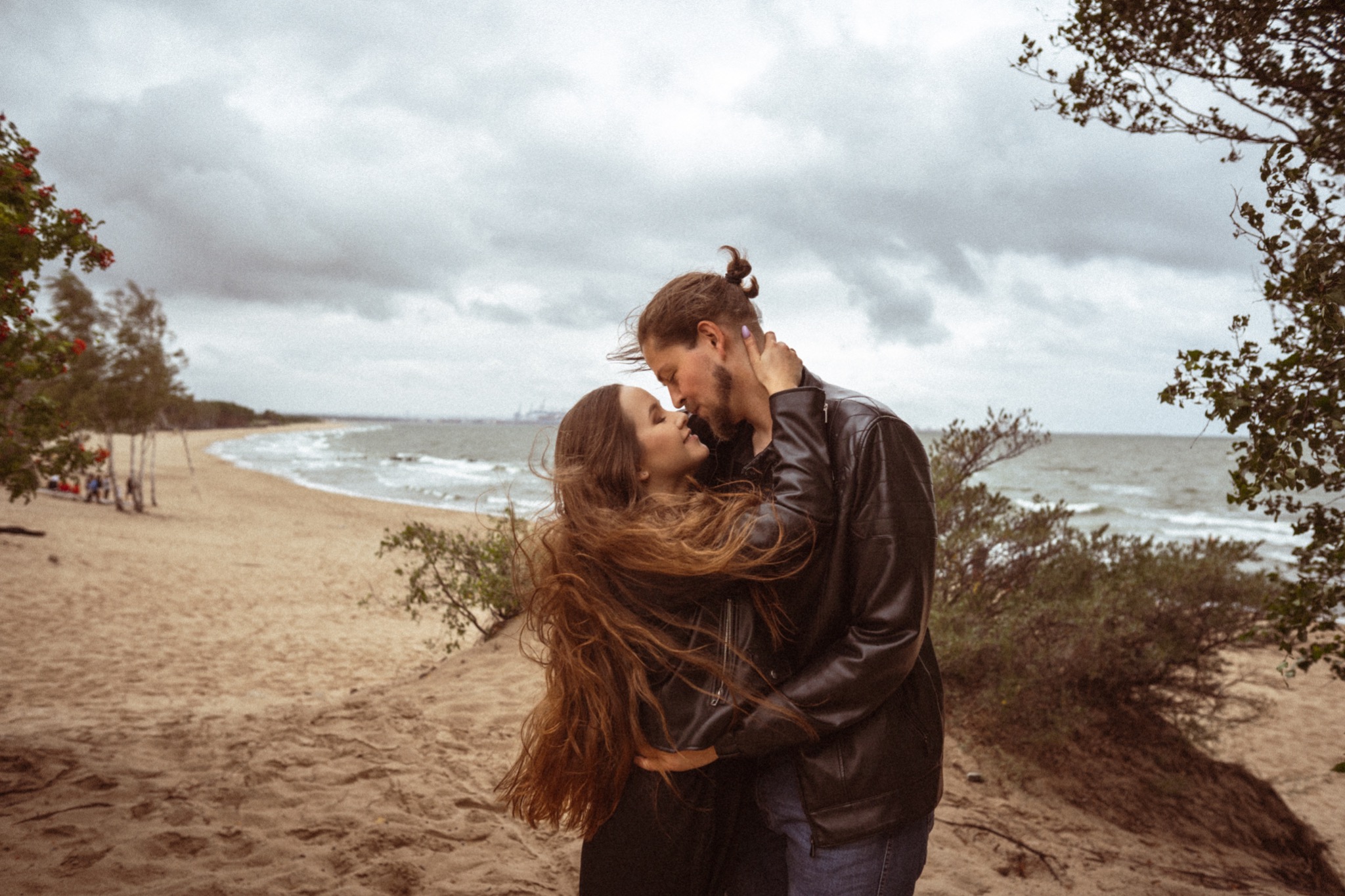 Para zakochanych, Ola i Mariusz, obejmują się na piaszczystej plaży nad morzem w pochmurny dzień. Ona z długimi, rozwianymi włosami, on w skórzanej kurtce.