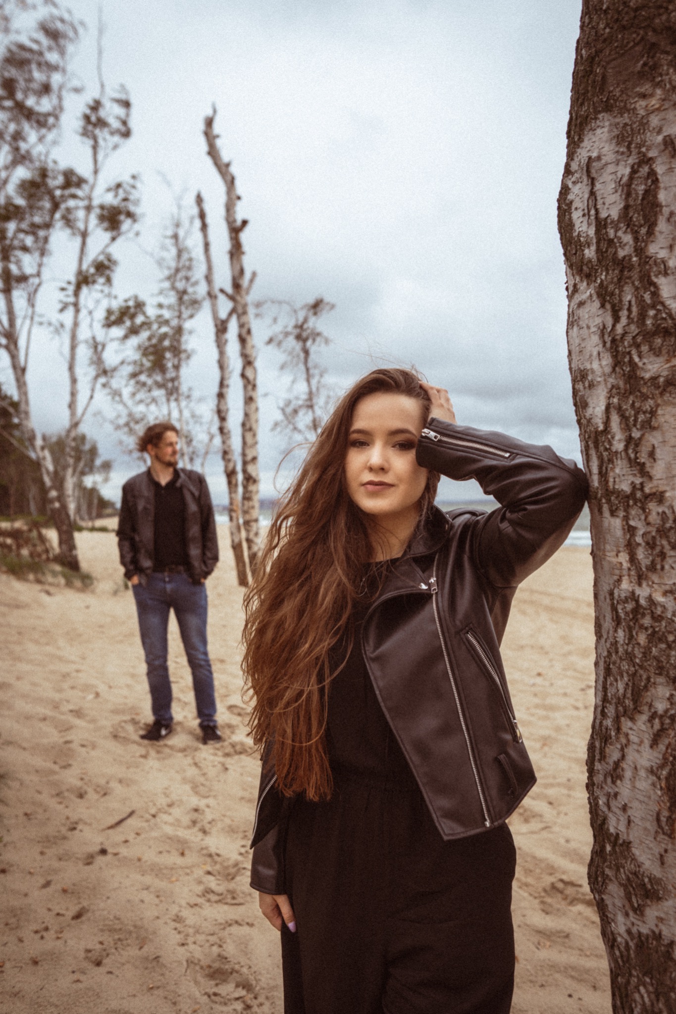 Para na plaży: kobieta w czarnej skórzanej kurtce opiera się o brzozę, mężczyzna w tle w dżinsach i kurtce, pochmurne niebo.