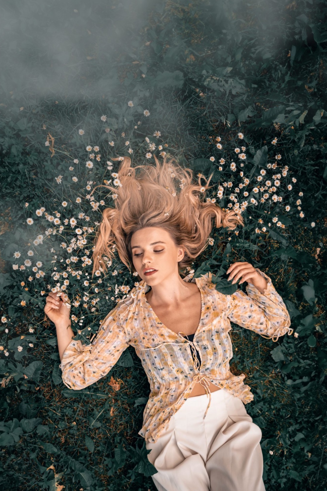 Kobieta w kwiecistej bluzce i białych spodniach leży na trawie wśród stokrotek, z zamkniętymi oczami, ujęcie z góry.