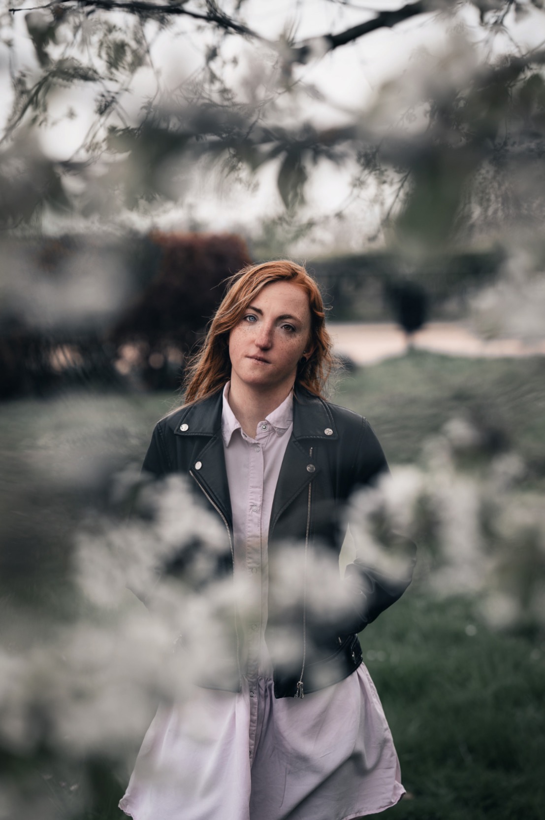 Portret kobiety z rudymi włosami i piegami, ubrana w skórzaną kurtkę i jasną sukienkę, pozująca na tle zieleni i kwitnących gałęzi.