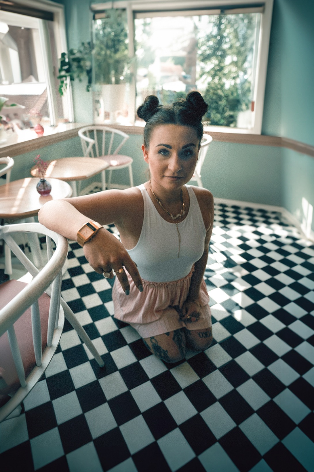 Kobieta w białym topie i różowej spódnicy klęczy na czarno-białej podłodze w kratkę, opierając się o białe krzesło, z włosami upiętymi w dwa koczki i złotą biżuterią, w jasnym wnętrzu kawiarni.