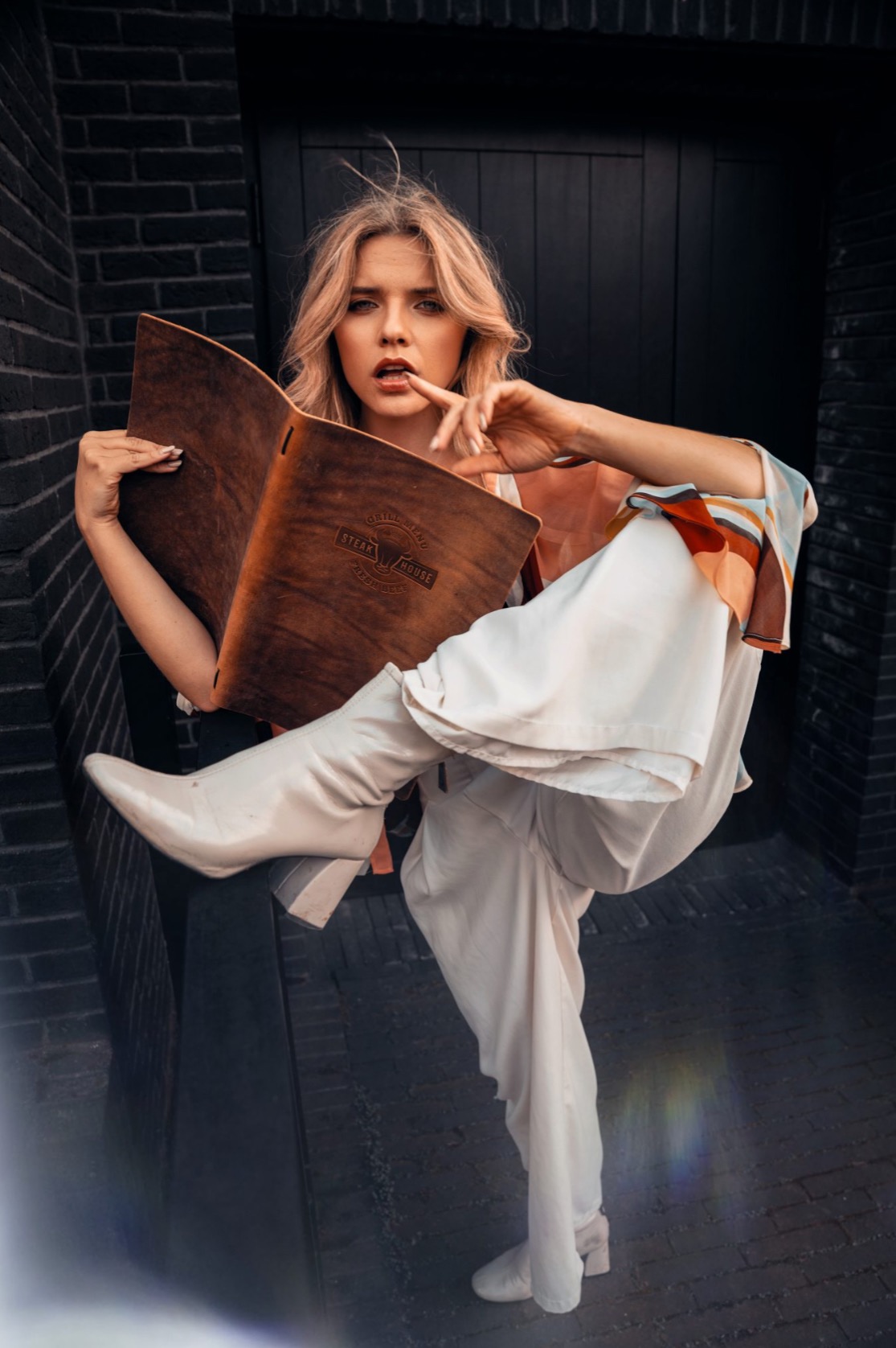 Blondwłosa kobieta w białym stroju i kremowych kozakach pozuje z drewnianym menu restauracji Steak House na tle czarnej ceglanej ściany.
