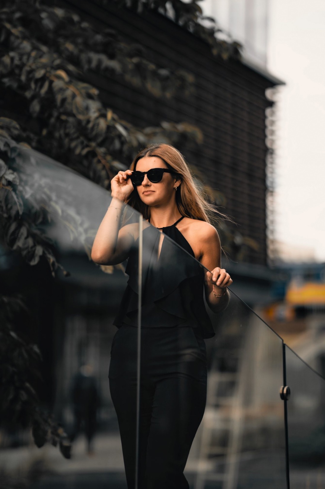 Elegancka blondynka w czarnym kombinezonie i okularach przeciwsłonecznych, pozująca na tle miejskiego krajobrazu z elementami zieleni i nowoczesnej architektury.