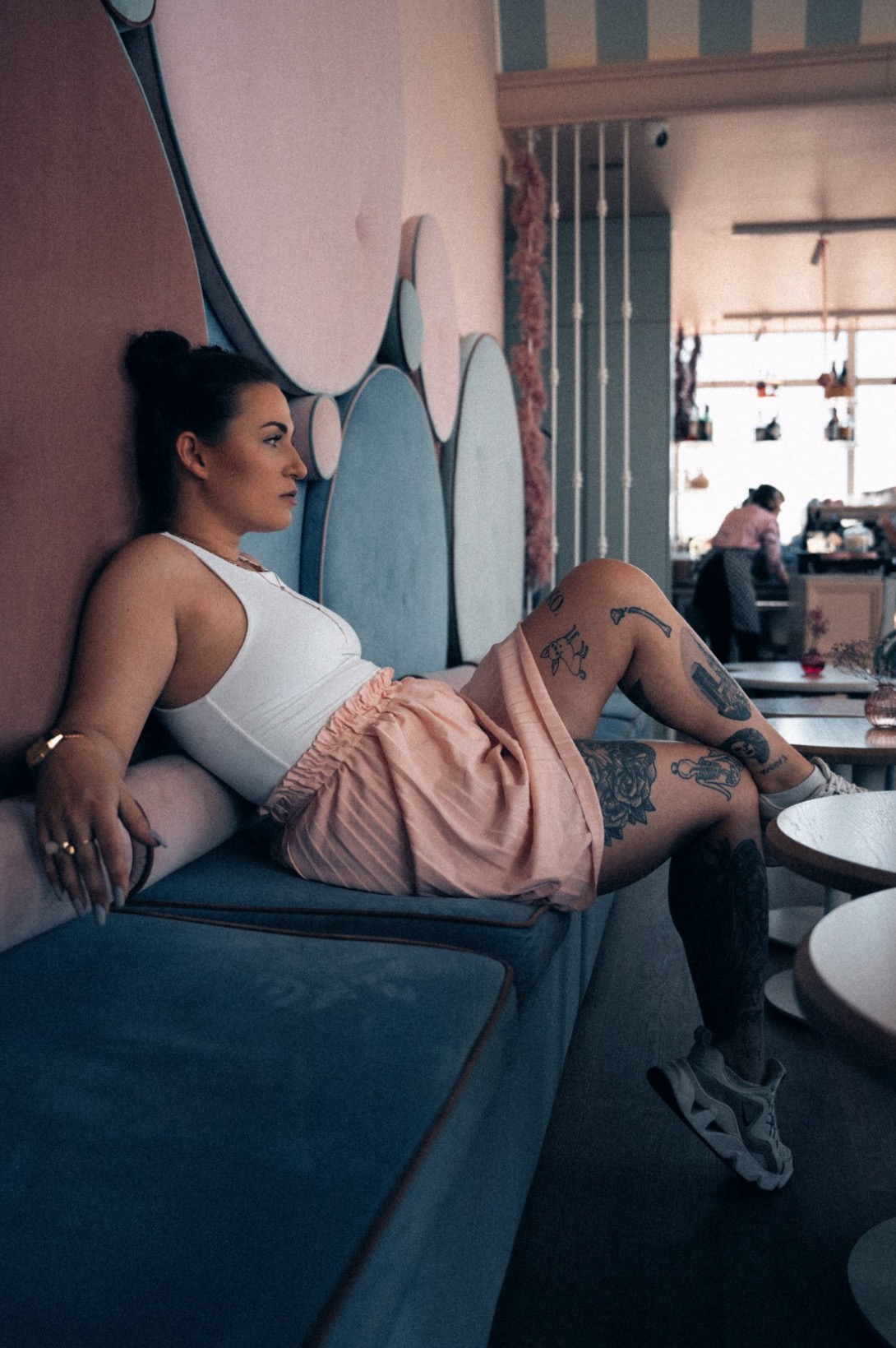 Kobieta z tatuażami na nogach, w białym topie i różowej spódniczce, siedzi na niebieskiej sofie z ozdobnym, kolorowym zagłówkiem w kawiarni, w tle widoczna osoba przy barze.