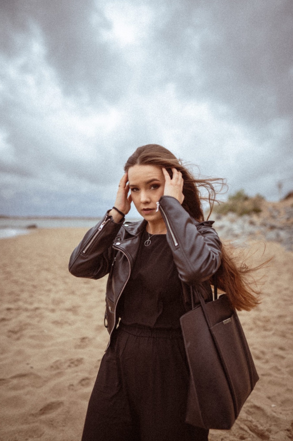 Portret młodej kobiety w czarnej skórzanej kurtce i sukience na plaży przy pochmurnym niebie, z rozwianymi włosami i torbą na ramieniu, patrzącej w obiektyw z rękami uniesionymi do głowy.