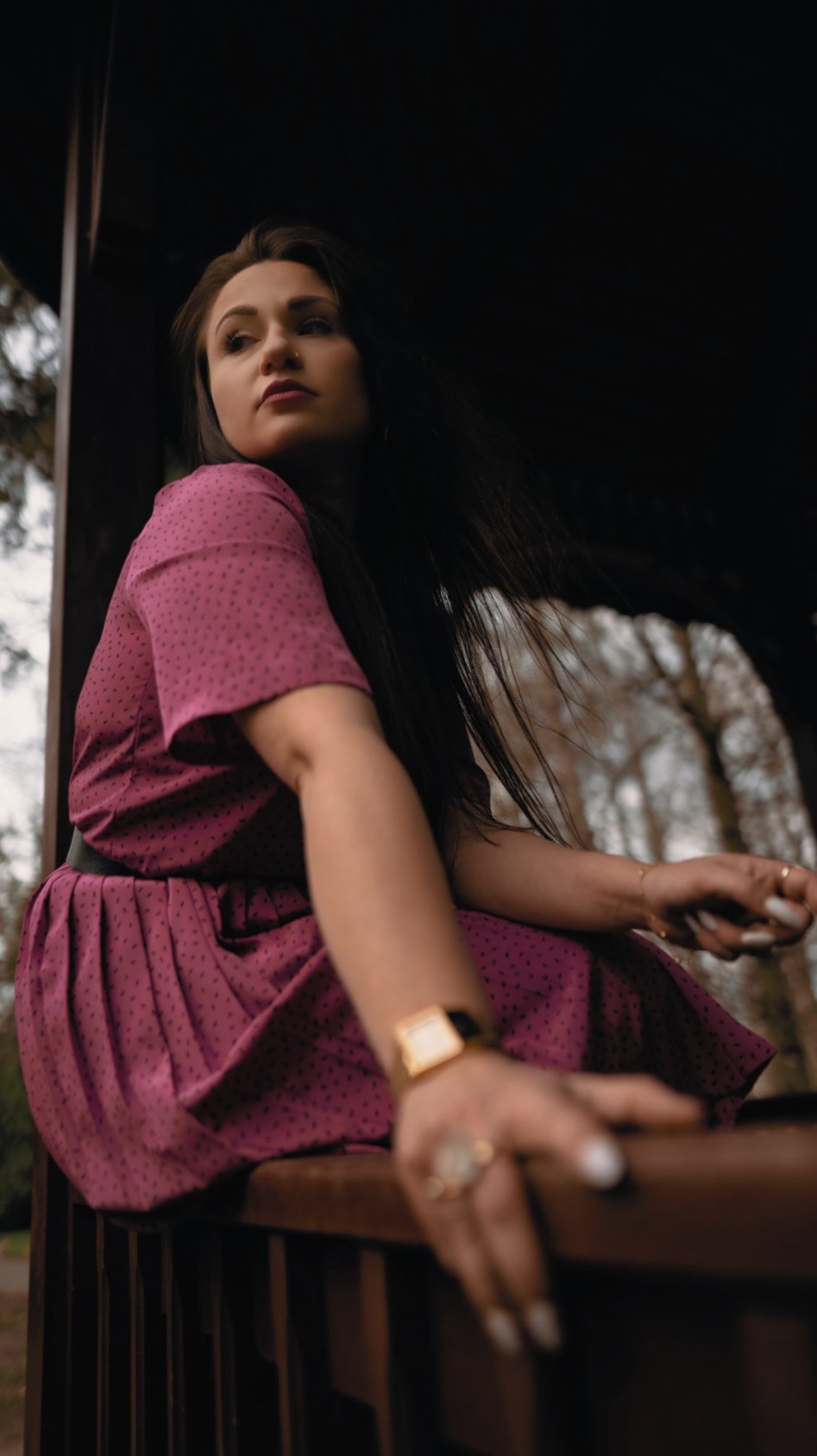 Kobieta w fioletowej sukience w kropki siedzi w altanie, spoglądając w lewo, z widocznym złotym zegarkiem i pierścionkiem na palcu.