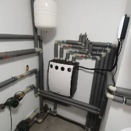 Biała pompa ciepła z czarnymi akcentami, zamontowana na ścianie w kotłowni, z widocznymi rurami instalacyjnymi, zbiornikiem wyrównawczym i manometrem, na szarych płytkach podłogowych.