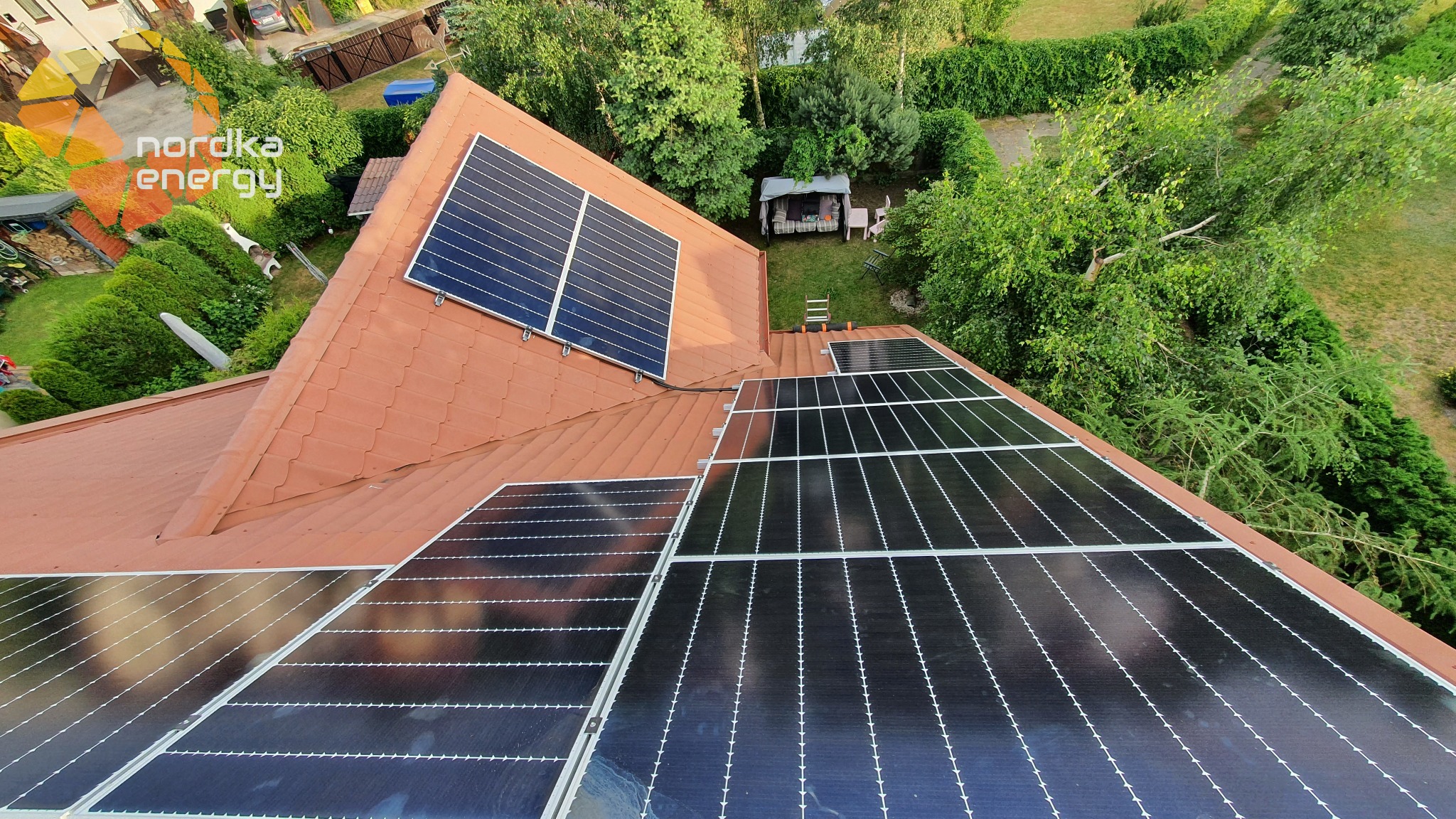 Panele fotowoltaiczne zamontowane na dachu domu jednorodzinnego z ogrodem, widok z góry, logo firmy w lewym górnym rogu.