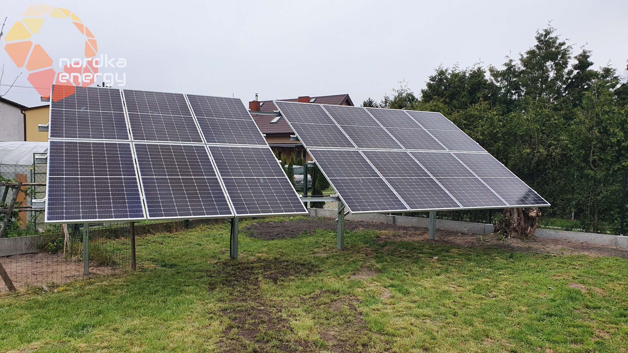 Dwa panele fotowoltaiczne ustawione na metalowych podporach na trawniku, w tle dom z czerwonym dachem i zielone drzewa, widoczny znak firmowy na panelu po lewej stronie.