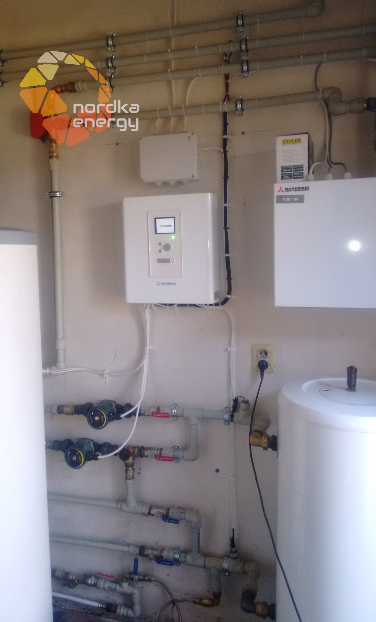 Montaż instalacji z białymi zbiornikami, rurami i urządzeniami Mitsubishi, widoczne pompy obiegowe i zawory, logo Nordka Energy w górnym rogu.