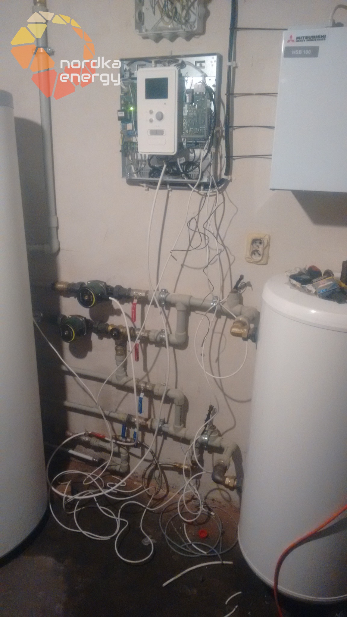 Węzeł cieplny z dwoma zbiornikami, widoczne orurowanie, zawory, sterownik z otwartą obudową, przewody i logo Nordka Energy w lewym górnym rogu.
