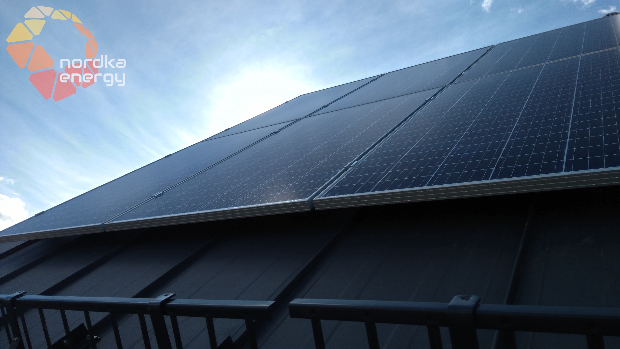 Panele fotowoltaiczne na dachu z blachy, widoczne z dołu pod kątem, logo firmy w lewym górnym rogu, błękitne niebo z chmurami w tle.