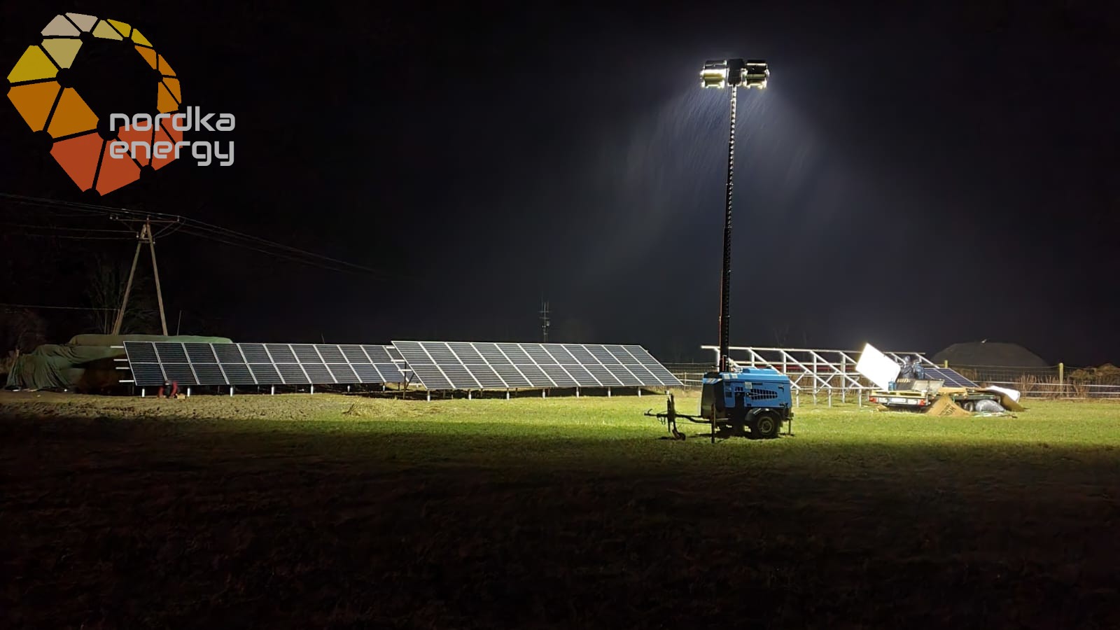 Instalacja paneli fotowoltaicznych oświetlona mobilnym masztem z reflektorami w nocy, widoczne panele, generator prądu na przyczepce i logo firmy Nordka Energy.