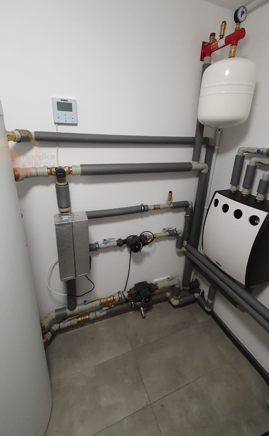 Węzeł cieplny w kotłowni: biały zbiornik wyrównawczy, czarno-biała obudowa urządzenia, izolowane rury, manometry i sterownik na ścianie, całość na szarych płytkach podłogowych.