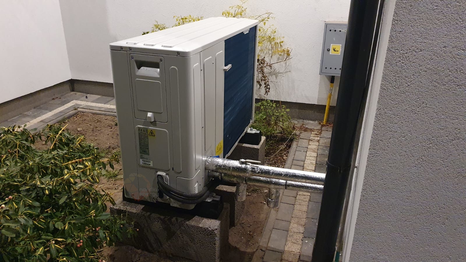 Zewnętrzna jednostka pompy ciepła zamontowana na betonowych bloczkach, z izolowanymi rurami, obok ściany budynku i skrzynki elektrycznej, otoczona roślinnością.