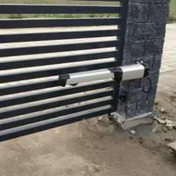 Automatyka do bram: Siłownik zamontowany na metalowej bramie posesji, widoczne narzędzia i materiały budowlane na ziemi, sugerujące trwający montaż.
