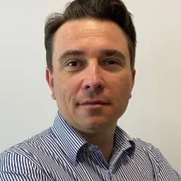 Portret mężczyzny w koszuli w paski, z lekkim uśmiechem i włosami zaczesanymi do tyłu, na neutralnym tle.