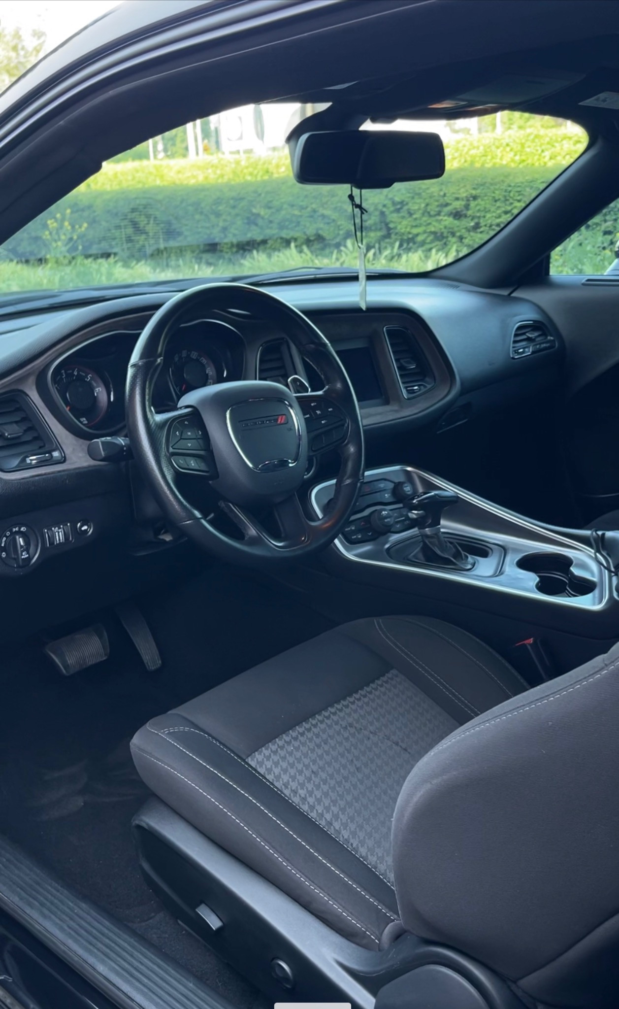 Wnętrze czarnego Dodge Challenger z widoczną deską rozdzielczą, kierownicą z logo i fotelem kierowcy z szarą tapicerką w pepitkę. Widok z zewnątrz przez szybę.