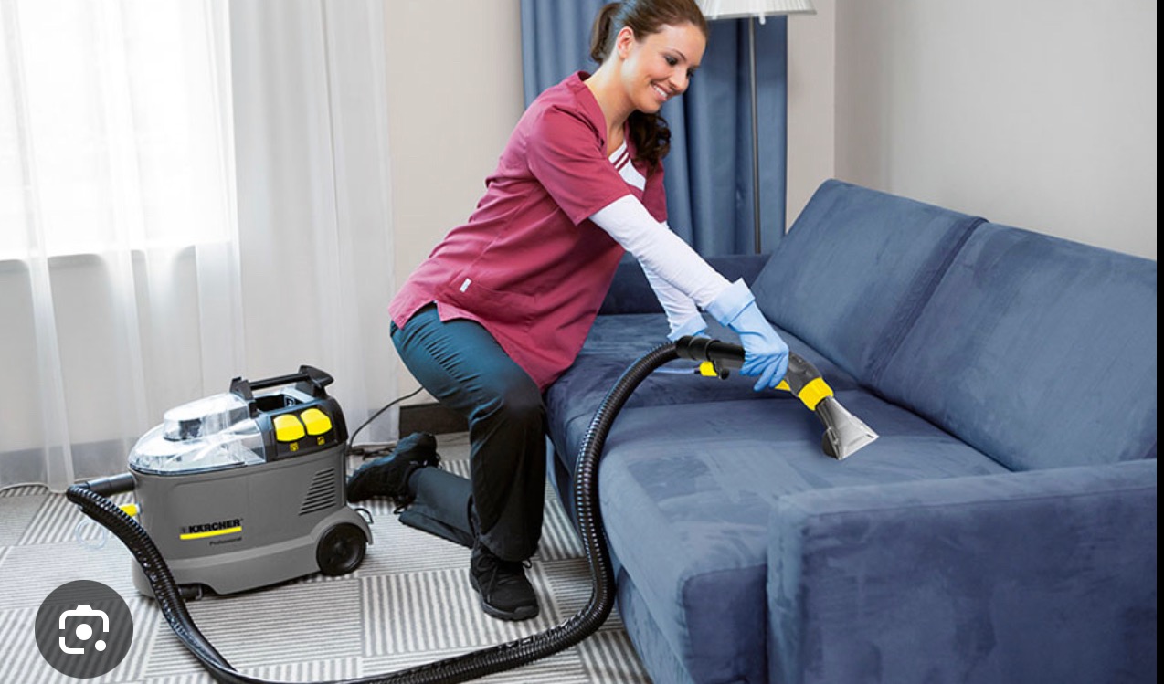 Kobieta w uniformie czyści niebieską kanapę odkurzaczem piorącym Karcher. Widoczny fragment okna z firanką i wzorzysta podłoga. Profesjonalne pranie tapicerki.