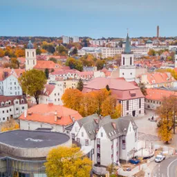 Panorama miasta z perspektywy lotu ptaka w jesiennej scenerii, z widocznymi wieżami kościołów, czerwonymi dachami budynków i placem budowy w tle.