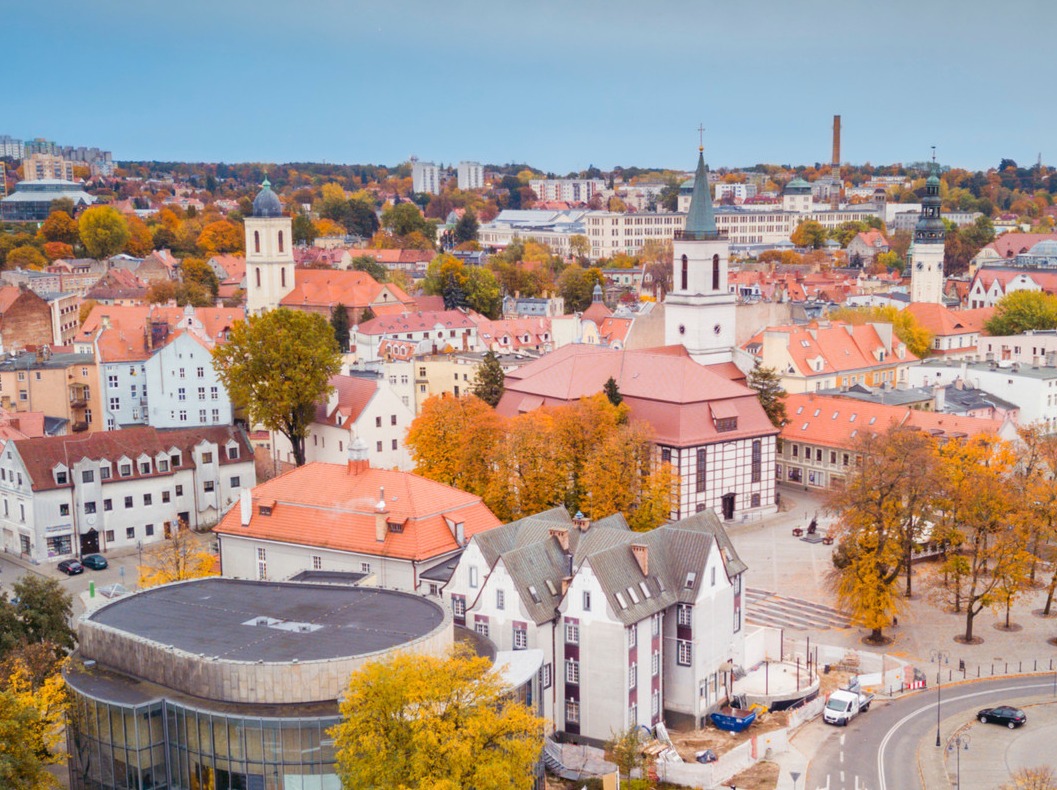 Panorama miasta z perspektywy lotu ptaka w jesiennej scenerii, z widocznymi wieżami kościołów, czerwonymi dachami budynków i placem budowy w tle.