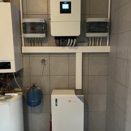 Kar-solar Usługi Hydrauliczne Karol Działa - Zestaw urządzeń: inwerter, rozdzielnice i akumulator energii Felicitysolar, zamontowane na ścianie z szarych płytek. Widoczne przewody i rury instalacyjne.