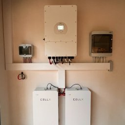 Kar-solar Usługi Hydrauliczne Karol Działa - Biały inwerter i dwa akumulatory CELLX zamontowane na ścianie wewnątrz pomieszczenia. Widoczne przewody i skrzynki elektryczne. Instalacja fotowoltaiczna w domu.