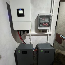 Kar-solar Usługi Hydrauliczne Karol Działa - Zamontowany inwerter z dwoma akumulatorami i rozdzielnicą na ścianie. Widoczne kable i rury instalacyjne. Szara kolorystyka akumulatorów.