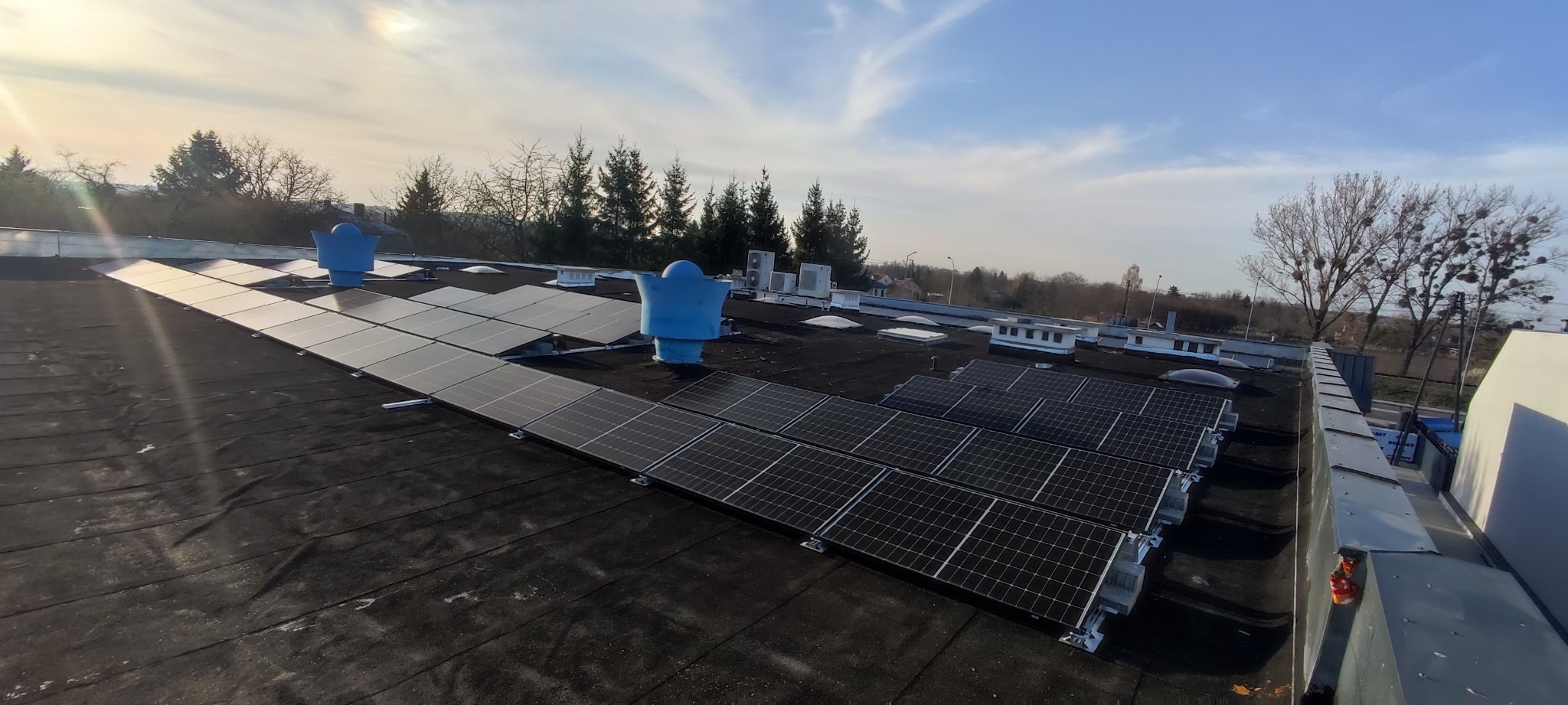 Instalacja fotowoltaiczna na płaskim dachu budynku przemysłowego, widoczne panele słoneczne w rzędach, kominki wentylacyjne oraz drzewa w tle pod błękitnym niebem z chmurami.