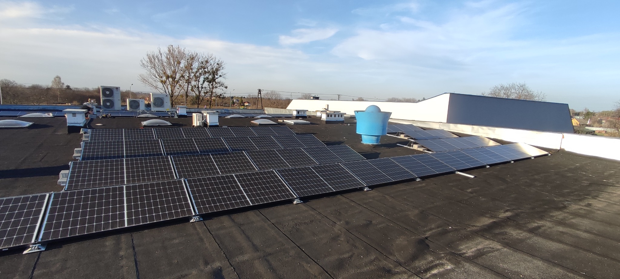 Panele fotowoltaiczne na płaskim dachu budynku przemysłowego, widoczne także klimatyzatory, świetliki dachowe i niebieski system wentylacyjny.